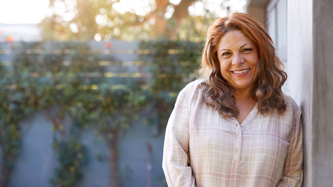 Portrait Of Smiling Senior Hispanic Woman In Garden At Home Against Flaring Sun